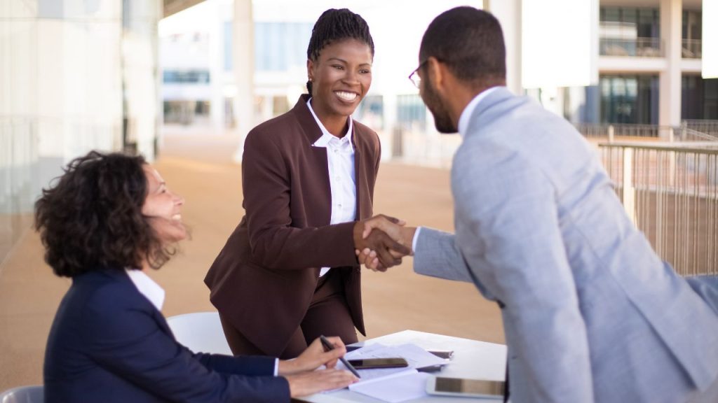 Three professionals meet outdoors; a woman in a brown suit stands and shakes hands with a man in a gray suit, nurturing client relationships, while another woman sits at a table with papers and a tablet, nurture your customers
