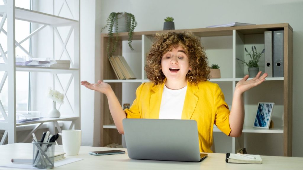 A person with curly hair and a yellow blazer sits at a desk with a laptop, raising hands in a questioning gesture. Their well-lit office space is filled with materials on nurturing your customers, suggesting they're pondering effective strategies for business growth, nurture your customers