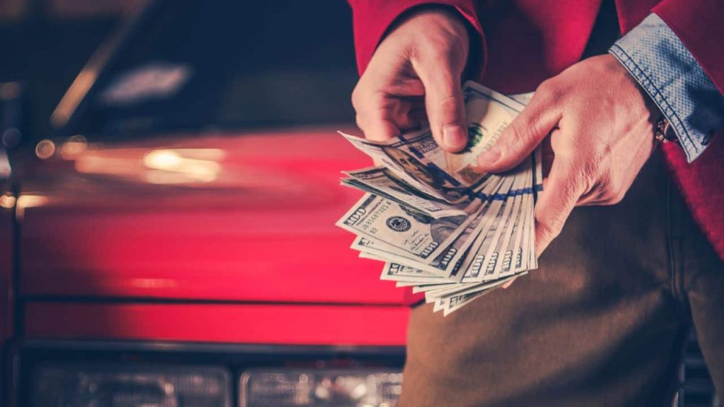 A person in a red jacket holds and counts a stack of U.S. hundred-dollar bills in front of a red car, symbolizing the excitement of generating new revenue streams, add new revenue streams