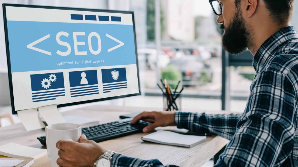 Advantages of Choosing a Local SEO Company - Top 6 Reasons A man in a plaid shirt works at a desk, viewing an SEO interface on a computer monitor, researching the advantages of choosing a local SEO company for his business.