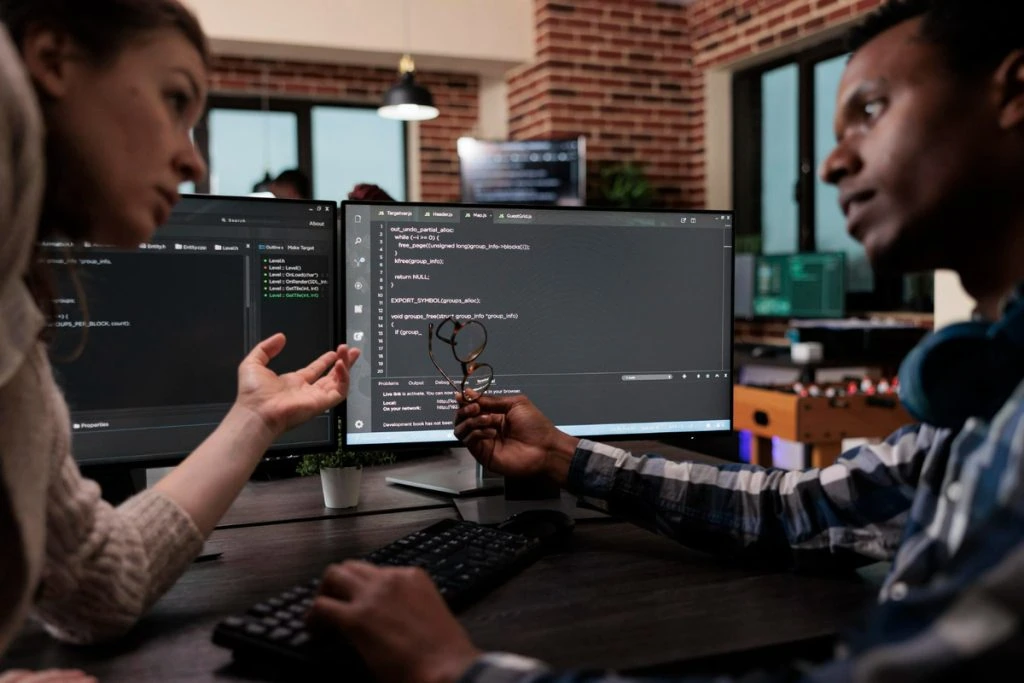 In an office with brick walls, two people engage in a discussion about WordPress website pricing. Dual monitors display a programming interface as one person holds eyeglasses and the other gestures animatedly with their hand.