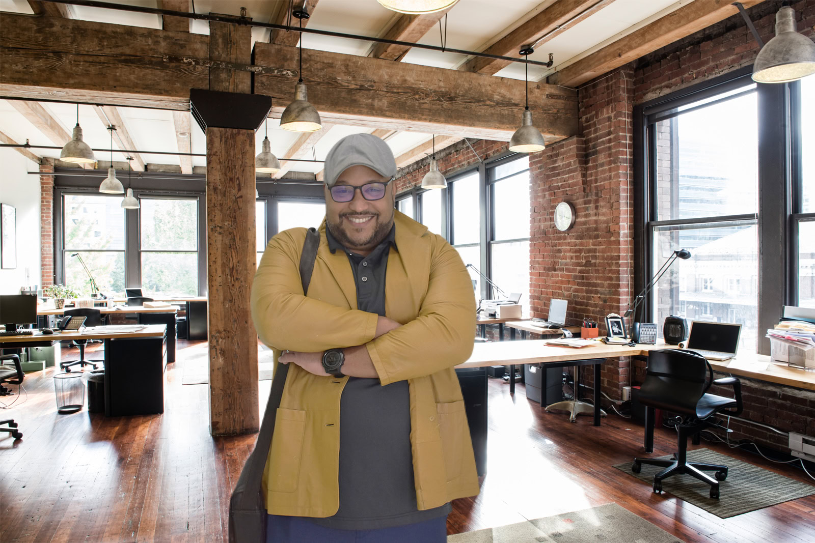 A man wearing glasses, a cap, and a yellow jacket stands smiling with arms crossed in a modern, open-plan office with large windows and exposed brick walls, where an ai call receptionist assists the team.